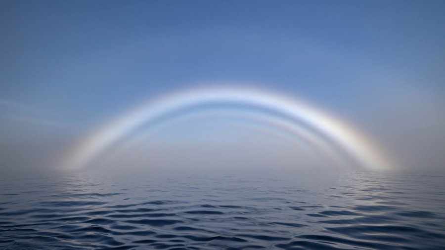 A very pale series of arch-like rainbows appears in fog above water.