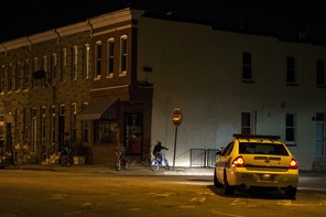 A police car sits on a street in East Baltimore, shining its headlights on a man riding a bike.