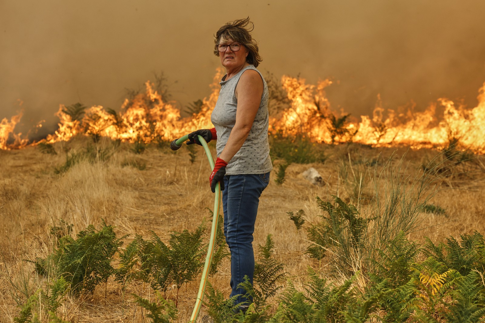A person holds a hose while standing in a field in front of an oncoming wildfire.
