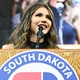 Kristi Noem stands at a podium at a 2023 rally in South Dakota and speaks into a microphone with a crowd behind her