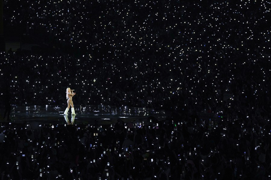 A performer sings onstage in a stadium, surrounded by many lights and phone screens held up by audience members.