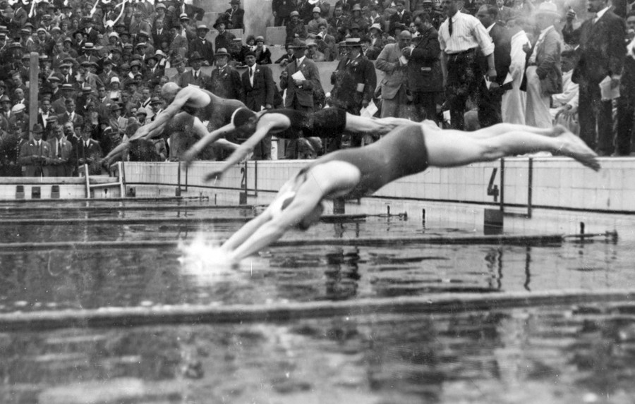 Swimmers dive in at the start of a race, watched by a large crowd.