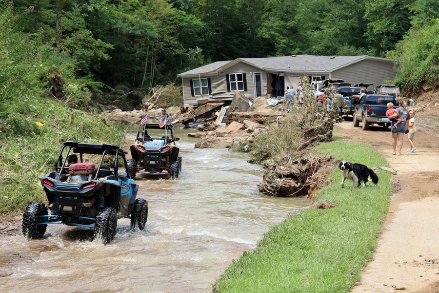 People work and drive near a damaged home that was pushed into a road by flooding.
