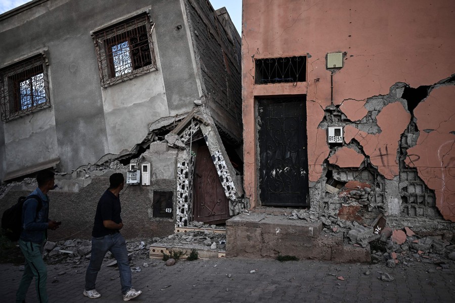 Two people walk past heavily damaged buildings.