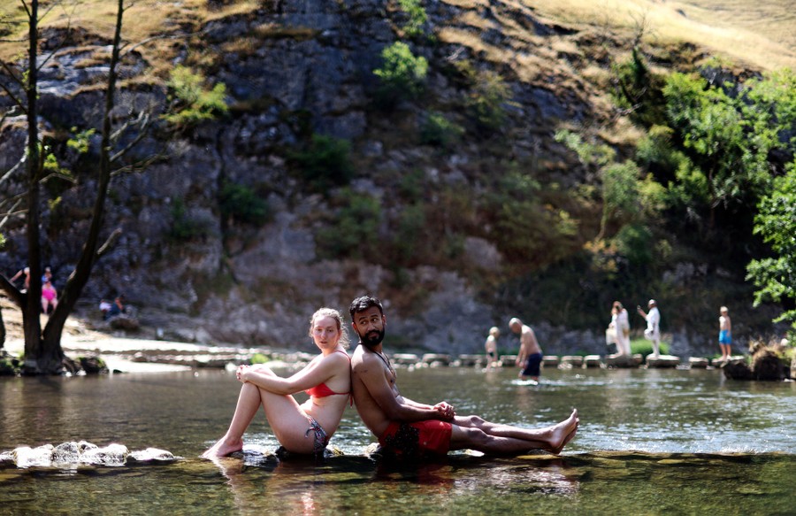 Two people sit back-to-back in a shallow river as others behind them play in the water.