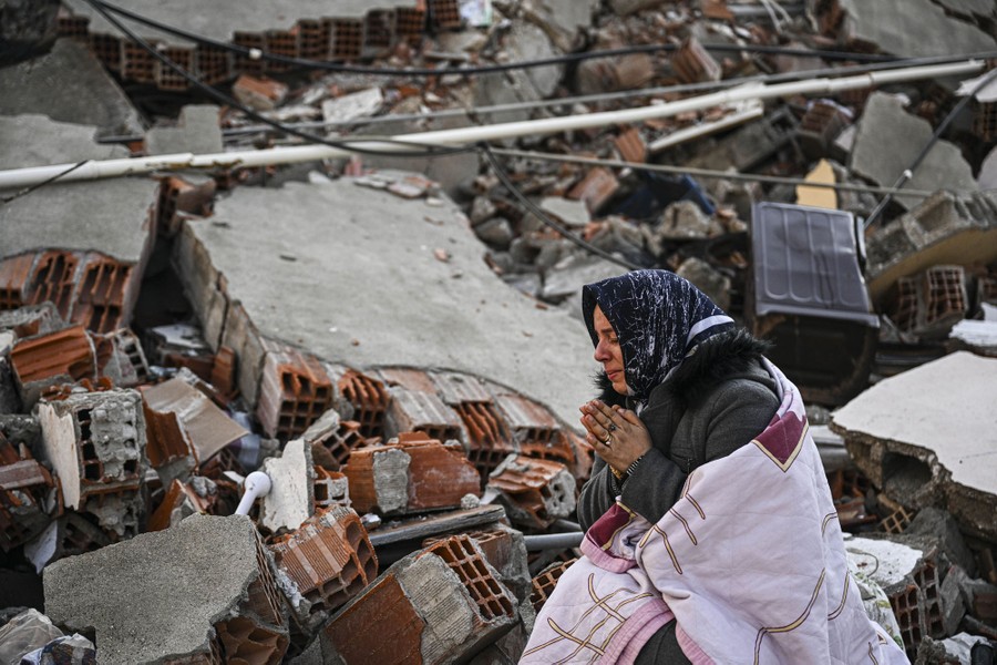 A woman, wrapped in a blanket, sits beside a large pile of rubble.