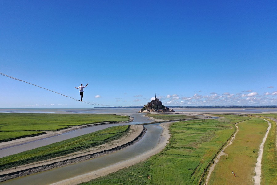 A person walks high on a slackline, above wetlands, toward a small rock outcrop with historical buildings crowded onto it.