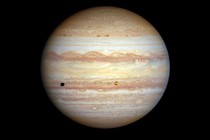 A view of the planet Jupiter, showing many broad and multi-colored bands of swirling storms in its atmosphere, and one small moon that casts a shadow