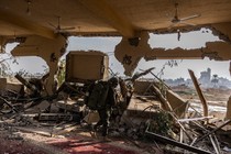 A soldier walks through a blown out interior in Gaza, ceiling fans and carpets visible under rubble but walls blasted through.