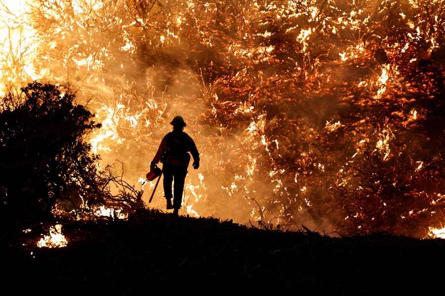 A firefighter walks in front of a hillside covered in burning brush.
