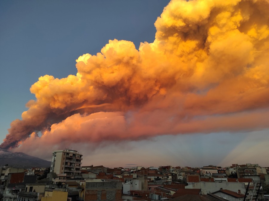Plumes of ash flow through the sky above buildings, at sunset.