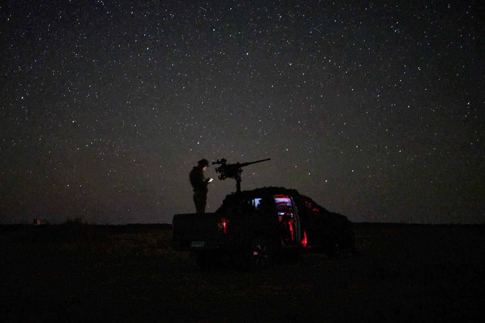 A soldier stands in a truck bed beside a mounted gun, seen under stars at night.