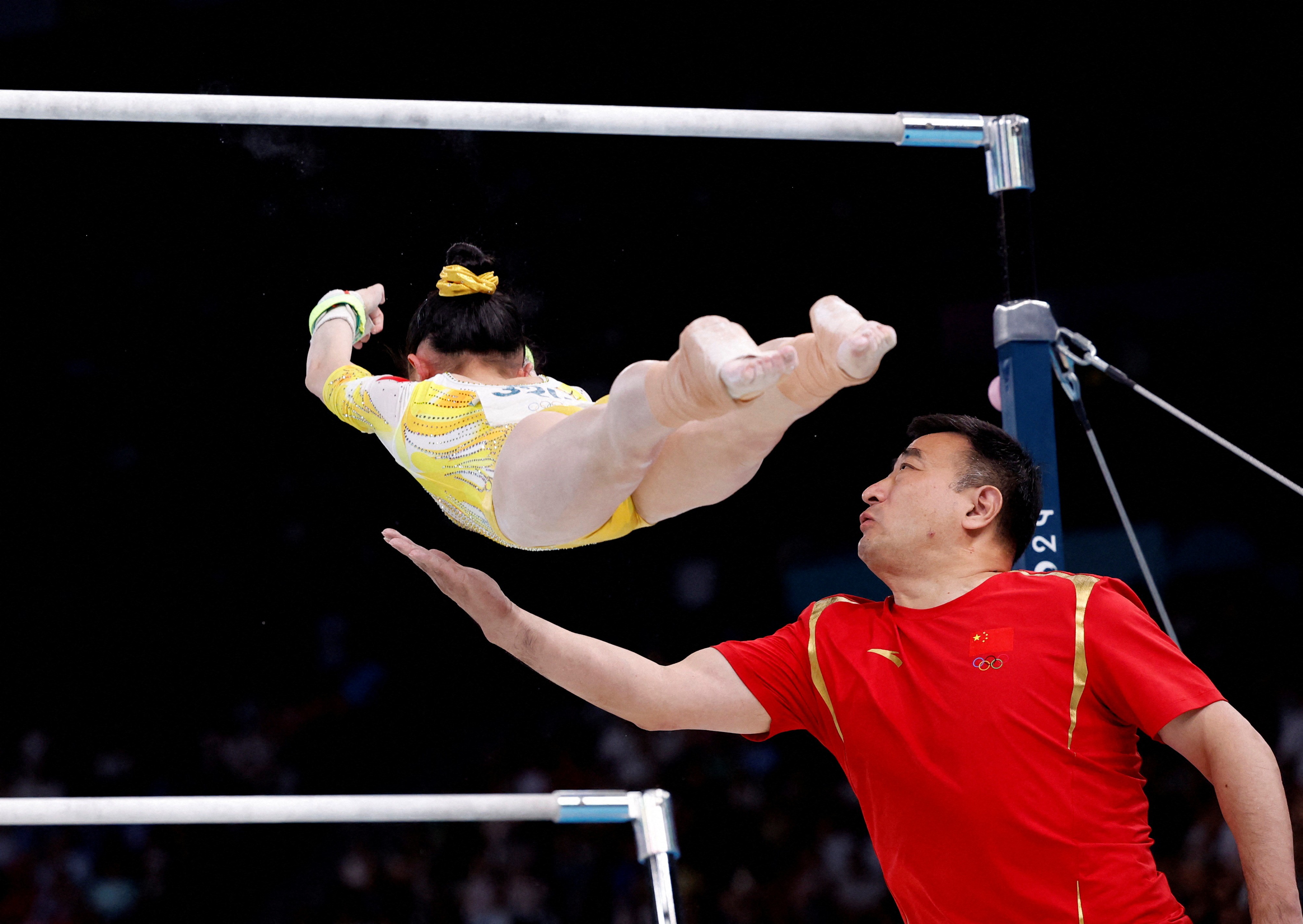 A gymnast falls face-down from a high bar she had been swinging from, as a coach reaches out one arm to slow her fall.