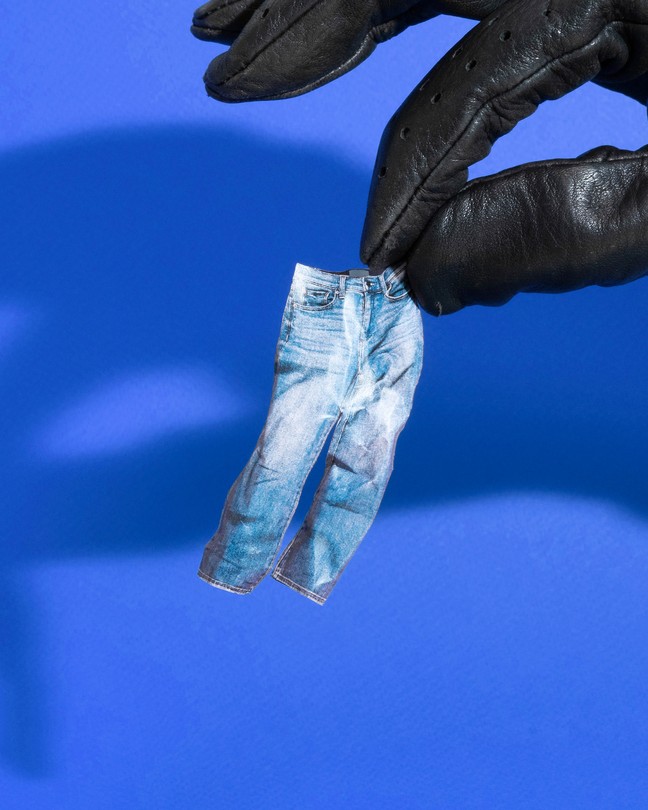 A black-gloved hand holding a miniature pair of crumpled paper jeans