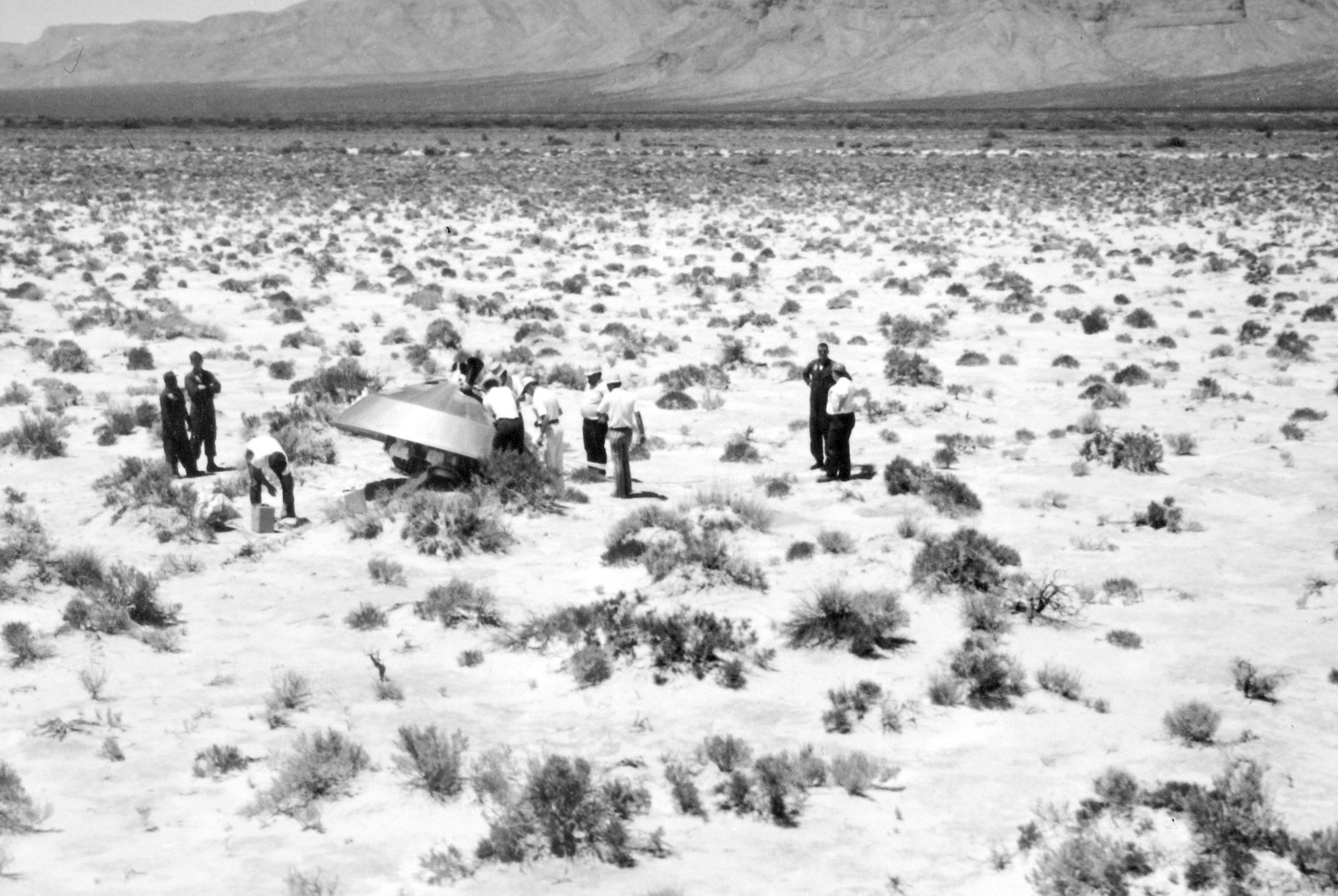 A black-and-white photograph of about a dozen people standing in a desert around a mock up of a spacecraft