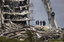 A half-dozen search-and-rescue team members stand atop a pile of rubble, beside what remains standing of a partially collapsed 12-story building.