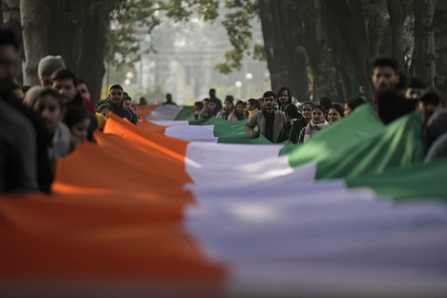People in a street hold up a very long sheet of fabric made of three colored stripes—one orange, one white, and one green—the flag of India.