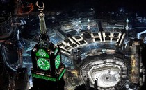 An aerial view of Mecca's Grand Mosque at night, featuring its huge, brightly lit clock tower.