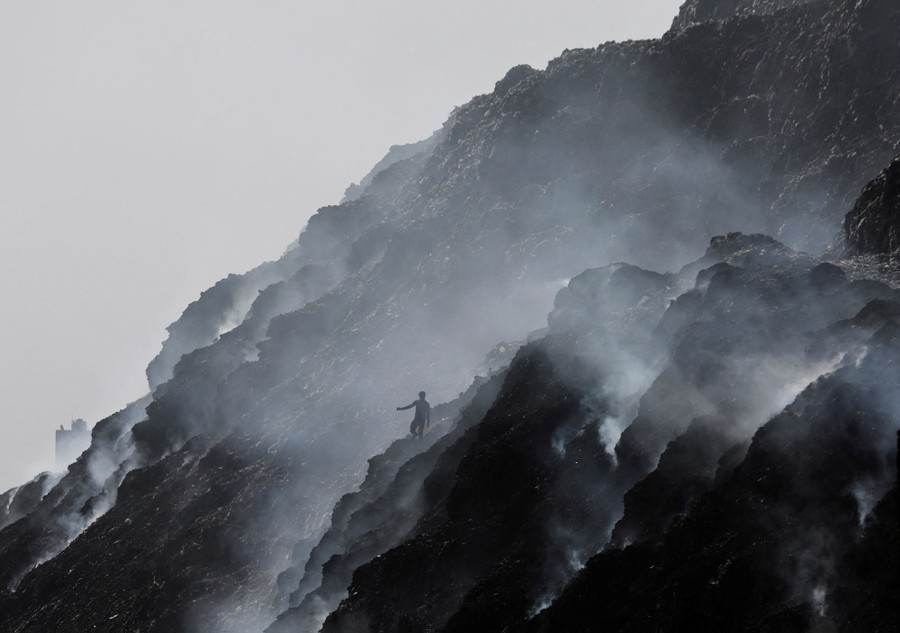 A person walks on an enormous smoking pile of garbage.