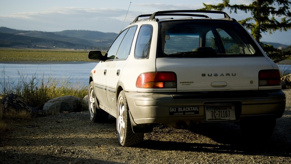 A white Subaru parked facing a body of water and mountains.