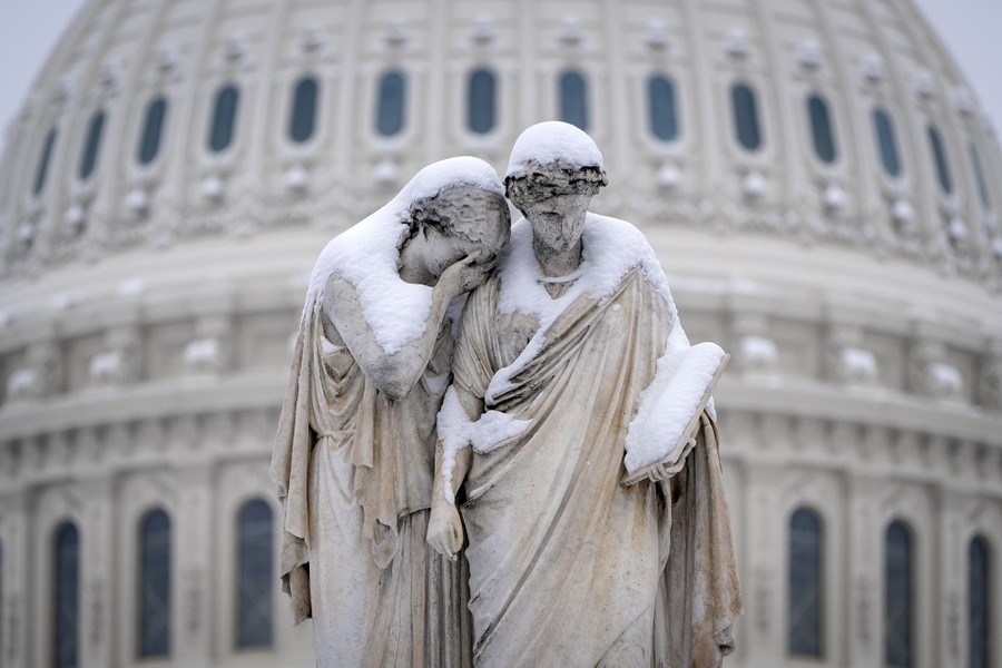 Statues of two figures standing side by side, covered in freshly fallen snow, in front of the U.S. Capitol building
