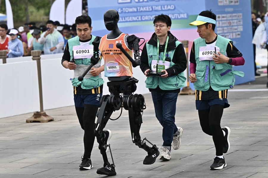 Several human minders trot along behind a humanoid robot as it runs in a race.