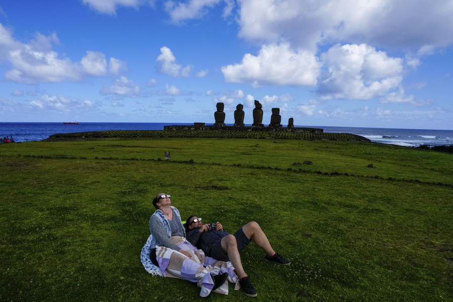 Two people lie on their backs on a field of grass, looking at the sky while wearing eclipse glasses, near several of the Moai statues on Easter island.