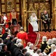 Queen Elizabeth and Prince Charles sit on golden thrones, surrounded by members of Parliament and other officials in both formal and official attire..