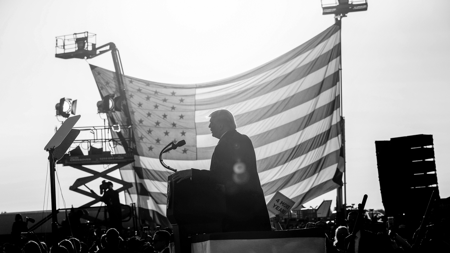 Black-and-white photo of Donald Trump giving a speech to a crowd in front of a large American flag suspended from two bucket lifts