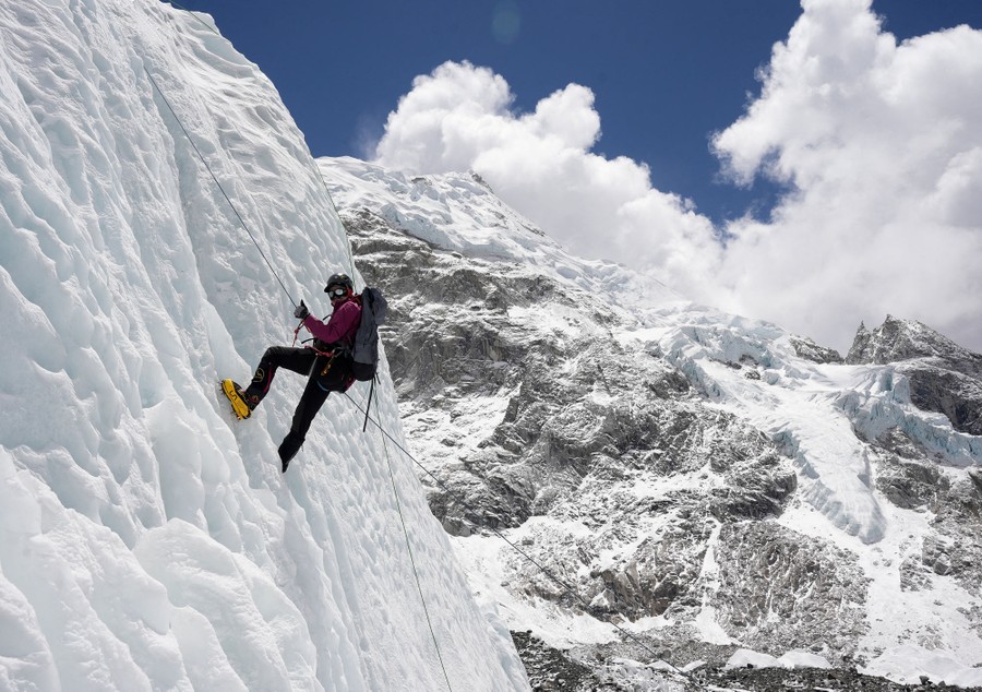 An ice climber hangs from a rope on the side of an ice cliff, with a tall mountain in the background.