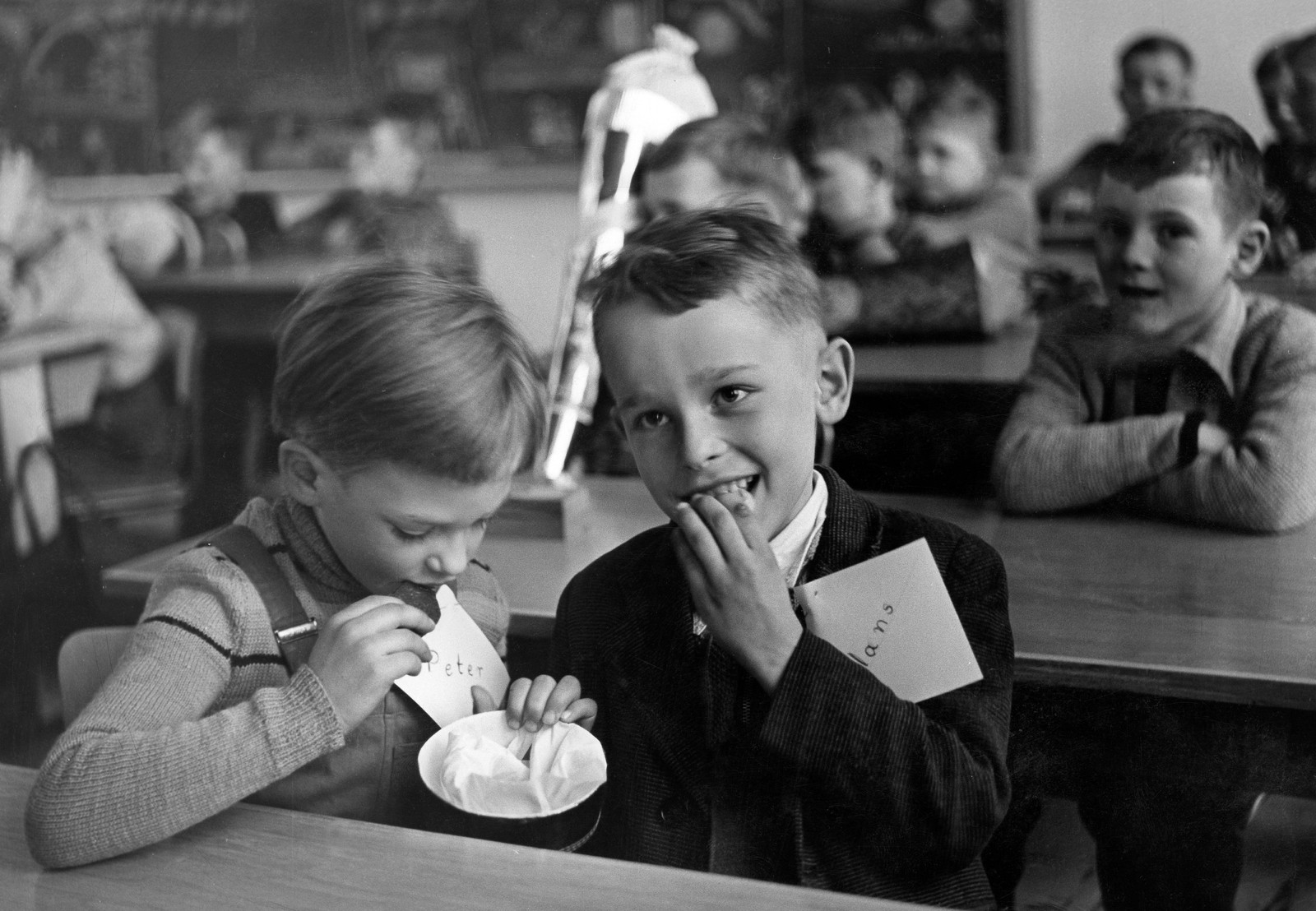 A black-and-white photograph of young children in a classroom, focusing on two boys who are sharing sweet treats.