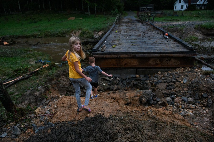 Two kids play on the remainder of a washed-out road, beside a small damaged bridge.