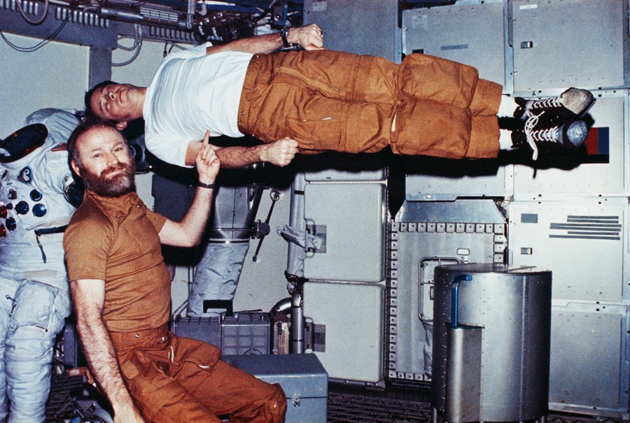 Inside a space station, one astronaut appears to hold another in the air with one finger as they both float in zero-g.