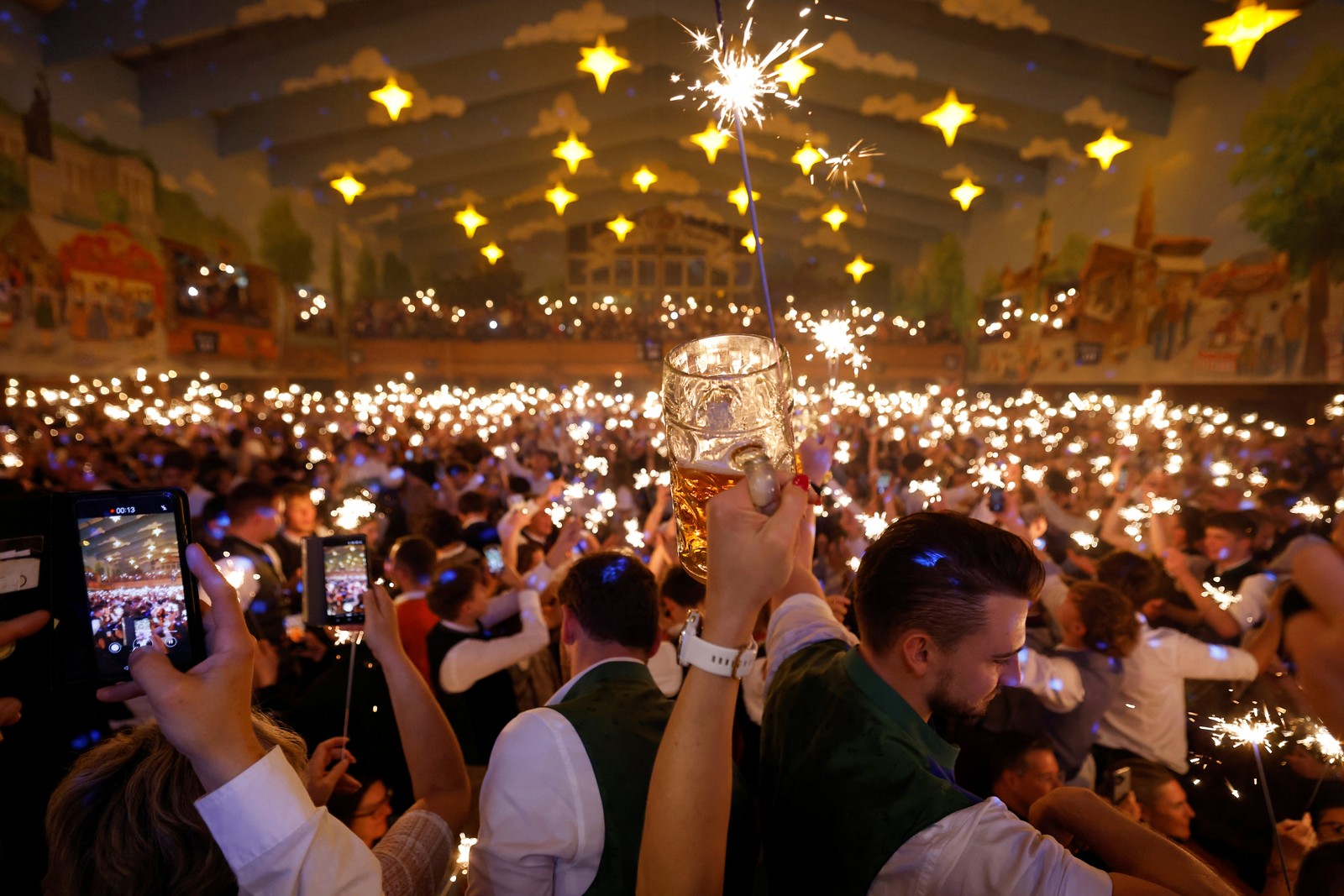A visitor raises a beer mug as a large crowd in a beer hall celebrates with sparklers.