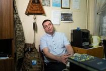 A man wearing a light-blue shirt sits in a chair against the wall in an office.