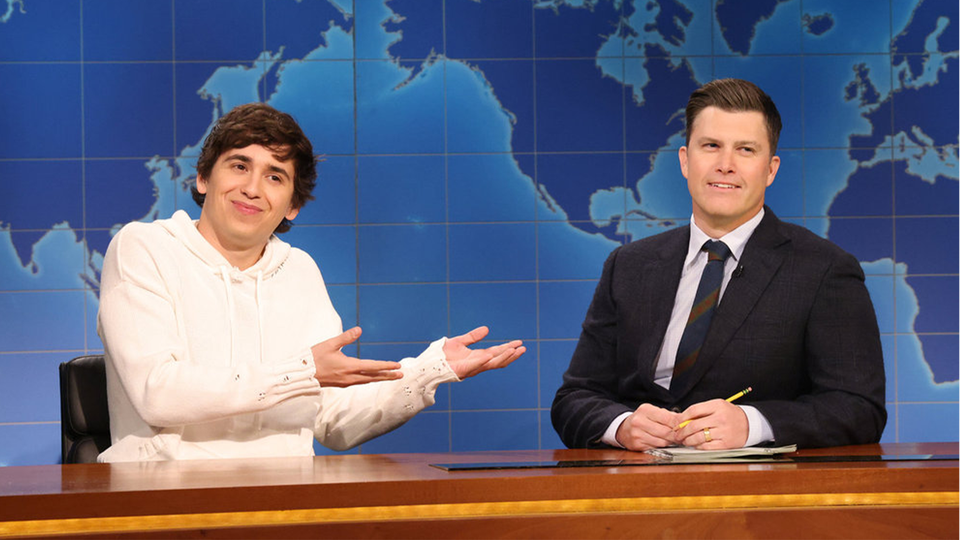 A man in a while hooded sweater points at a man in a suit with an image of a world map behind them.