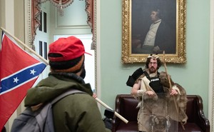 man dressed in pelts stands in capitol while a man with confederate flag looks on