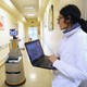 A human caregiver uses a laptop to operate a robot caregiver in the hallway of a nursing residence.