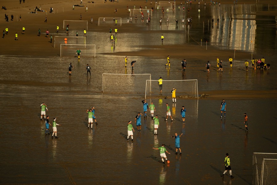 Many soccer players take part in matches on a broad, flat beach at low tide, across many temporary pitches.