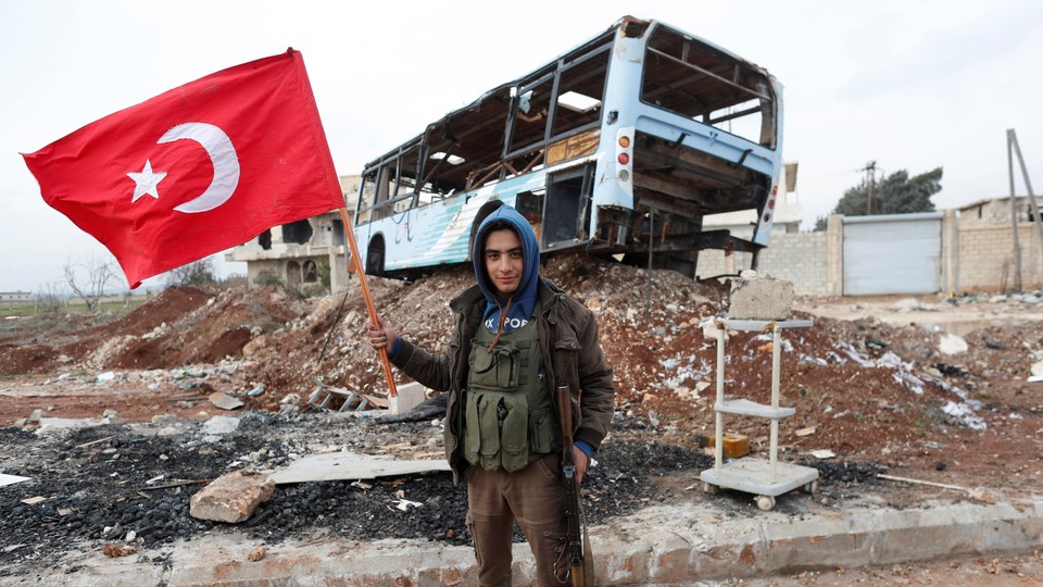 A Free Syrian Army fighter stands in front of a destroyed bus, holding a gun in one hand and a Turkish flag in the other.