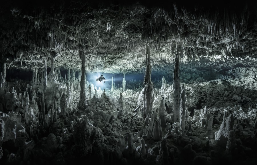 A diver swims through an underwater cave, lit by powerful flashlights.