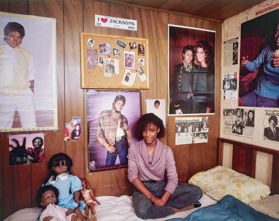 photo of girl in pink cowl-neck sweater and jeans sitting cross-legged on bed leaning against wood-paneled wall with many posters of Michael Jackson, a bulletin board, newspaper clippings, and a bumper sticker with "I (heart) JACKSONS"