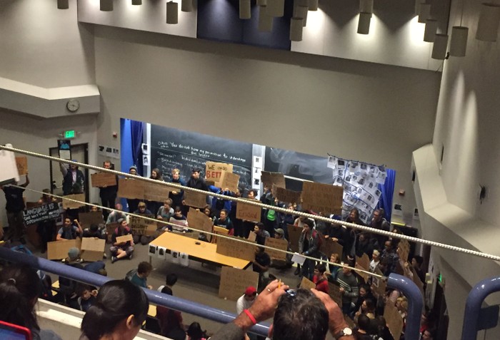 Students stand at the front of a lecture hall, holding signs.