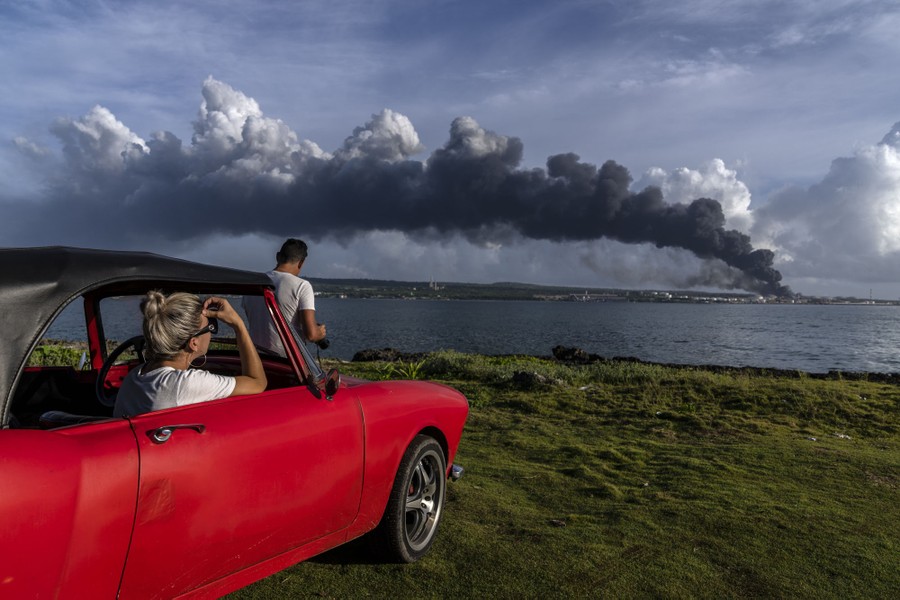 People in and on a parked car watch a plume of smoke rise from a site across a body of water.