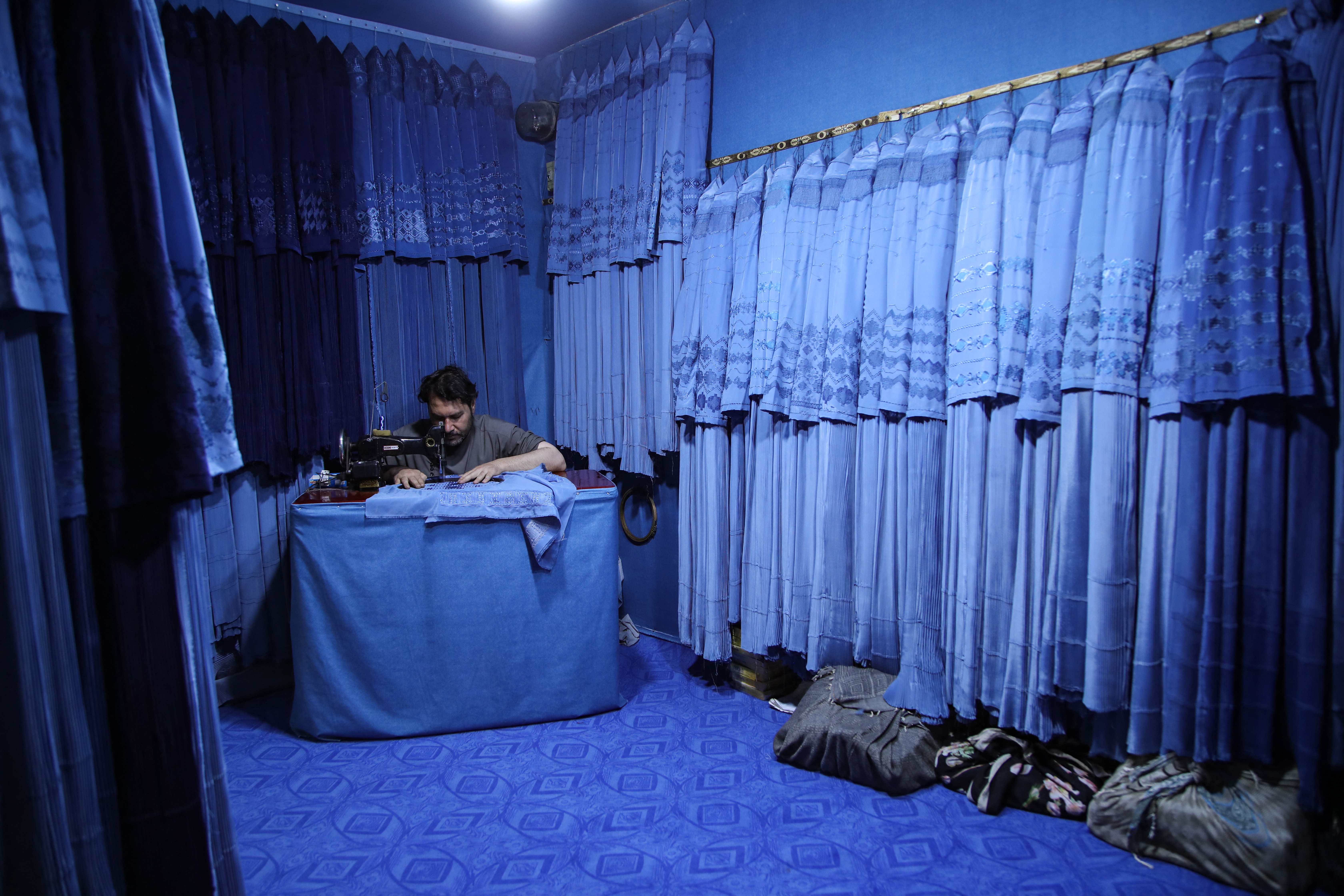 A tailor stitches burqas inside a shop surrounded by many other blue burqas.