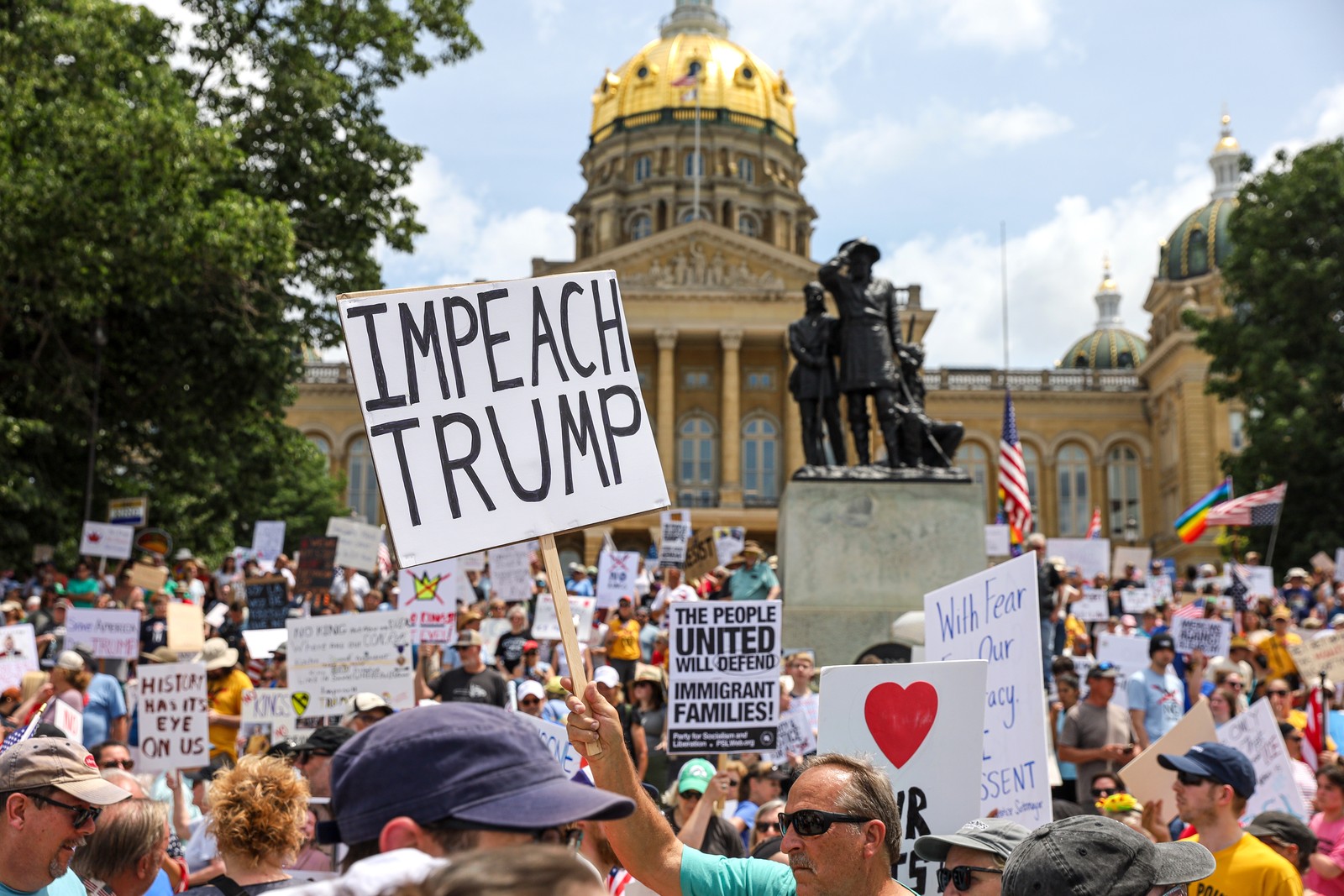 A crowd of protesters gathers outside a state capitol building carrying signs, including one which reads 