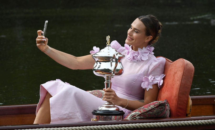 A tennis player wearing a dress sits in a small boat, holding a trophy, taking a selfie.