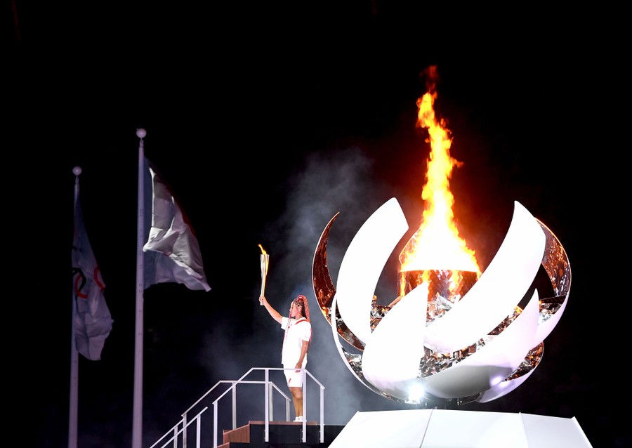 An athlete stands with a lit torch, while a large stylized cauldron behind them burns.