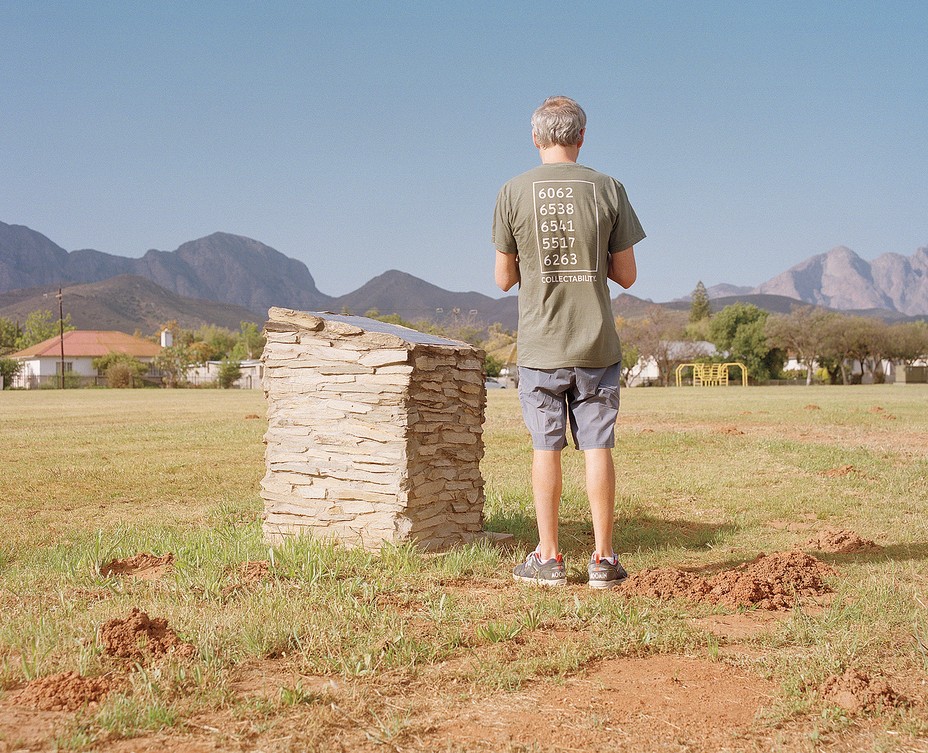 photo of author facing away from camera, wearing t-shirt and shorts, standing on grass in park next to stone marker with metal plaque on top, with playground, homes, and mountain range in background