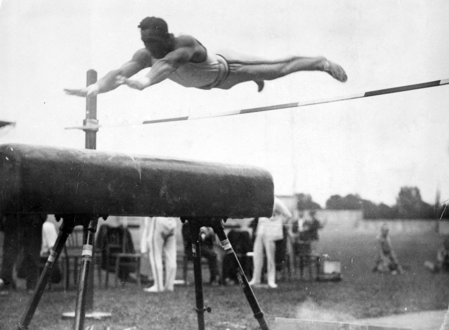 A gymnast is seen in mid-air, leaping over a bar toward a vaulting horse apparatus.